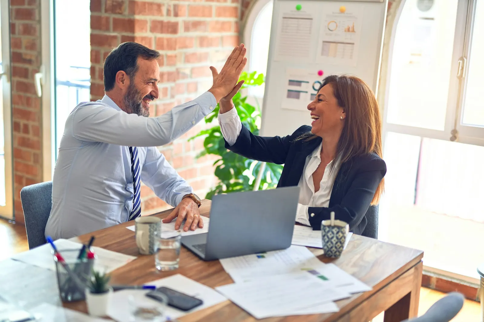people high fiving sitting at table with computer and paper(s)